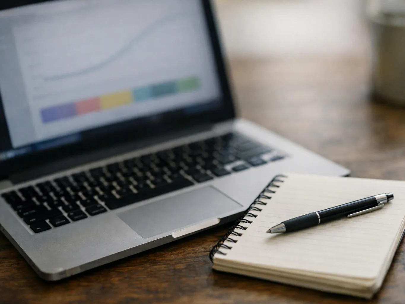 A laptop shows a blurred chart beside a spiral notebook and pen on a wooden table