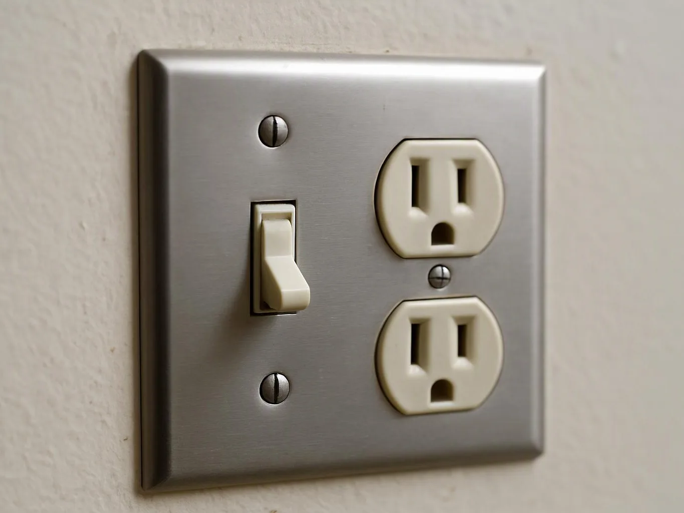 A wall switch and two electrical outlets sit on a brushed metal plate against a light wall
