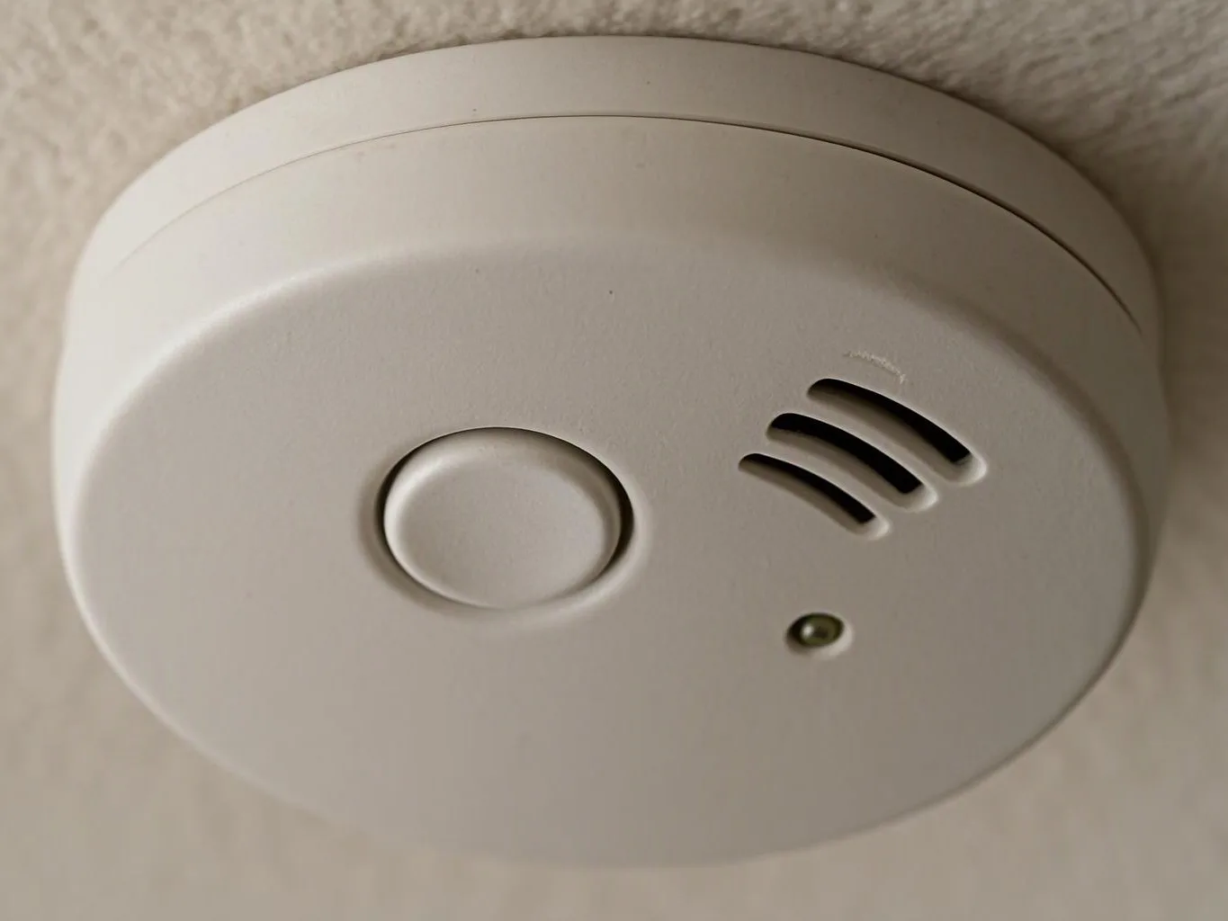 A white smoke alarm sits on a ceiling, with a test button, vents, and a small indicator light